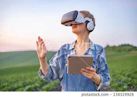 A person wearing a virtual reality headset and holding a tablet stands in a green field, exploring modern agricultural technology 138329926