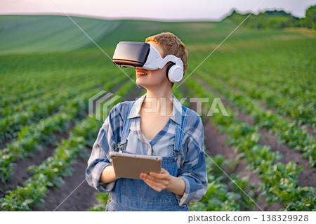 A young woman in overalls and a plaid shirt uses a tablet and VR headset while standing in a lush green field with rows of crops 138329928