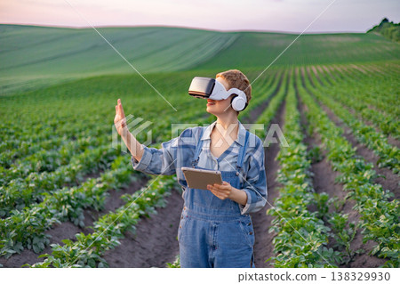 A person wearing a virtual reality headset and holding a tablet stands in a field of crops, exploring modern agricultural technology A person wearing a virtual reality headset and holding a tablet stands in a field of crops, exploring modern agricultural technology 138329930