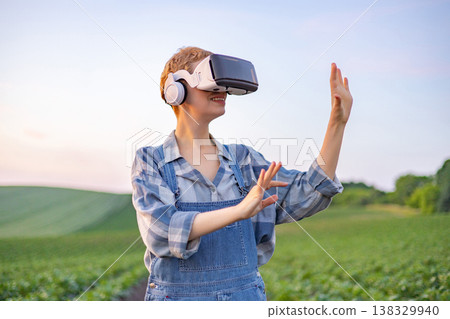 A young woman wearing overalls and a plaid shirt uses a virtual reality headset and headphones while standing in a lush green agricultural field at sunset A young woman wearing overalls and a plaid shirt uses a virtual reality headset and headphones while standing in a lush green agricultural field at sunset 138329940