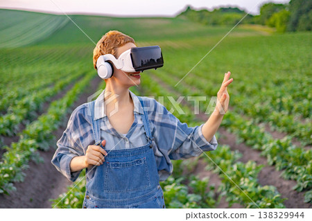 A young woman wearing a virtual reality headset and overalls stands in a lush green field, interacting with technology A young woman wearing a virtual reality headset and overalls stands in a lush green field, interacting with technology 138329944