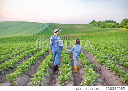 A woman in overalls and a child walk hand-in-hand through rows of green crops on a sunny day A woman in overalls and a child walk hand-in-hand through rows of green crops on a sunny day 138329965