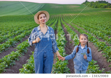 A woman and a young girl wearing denim overalls and straw hats stand hand-in-hand in a lush green field of crops A woman and a young girl wearing denim overalls and straw hats stand hand-in-hand in a lush green field of crops 138329969