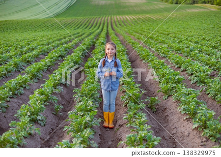 A young girl stands in the middle of a vast potato field, holding a small plant, surrounded by rows of green crops stretching to the horizon 138329975