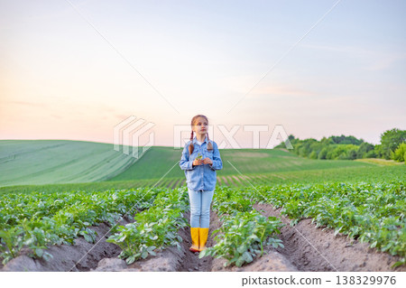 A young girl with braided hair stands in a lush potato field wearing a denim shirt and yellow boots, holding a small plant A young girl with braided hair stands in a lush potato field wearing a denim shirt and yellow boots, holding a small plant 138329976