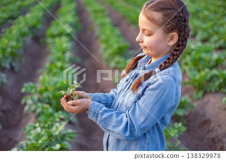 A young girl with braided hair carefully holds a small plant seedling in her hands while standing in a field of crops A young girl with braided hair carefully holds a small plant seedling in her hands while standing in a field of crops 138329978