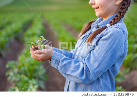 A young girl with braided hair holds a small green seedling in her cupped hands while standing in a cultivated field with rows of crops 138329979
