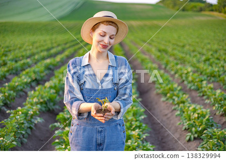 A woman in overalls and a straw hat stands in a field, holding a small seedling in her hands with care and pride 138329994