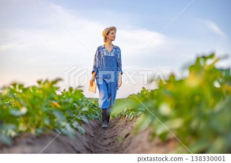 A woman in overalls and a straw hat walks through a field of crops holding a tablet device, looking off into the distance A woman in overalls and a straw hat walks through a field of crops holding a tablet device, looking off into the distance 138330001