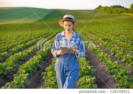 A young woman in overalls and a hat stands in a lush green field, examining data on a tablet computer A young woman in overalls and a hat stands in a lush green field, examining data on a tablet computer 138330021