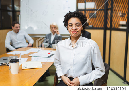 Portrait of young female team leader businesswoman in office, colleagues meeting in background. Confident African American woman business leader sitting in office. Boss leader professional worker 138330104