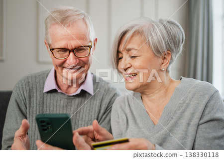Senior couple man woman shopping online with smartphone paying with credit card. Old people buying on Internet enter credit card details at home indoors. Online shopping delivery service ecommerce Senior couple man woman shopping online with smartphone paying with credit card. Old people buying on Internet enter credit card details at home indoors. Online shopping delivery service ecommerce 138330118