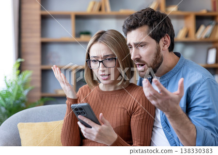 Young couple in a living room reacting with shock and frustration to unexpected bad news on a smartphone screen, tense and confused while scrolling and discussing the issue Young couple in a living room reacting with shock and frustration to unexpected bad news on a smartphone screen, tense and confused while scrolling and discussing the issue 138330383
