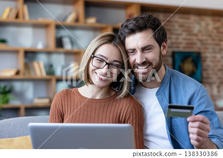 Smiling couple in their living room purchasing goods online, holding a credit card and looking at a laptop together, representing modern e-commerce and digital finance 138330386