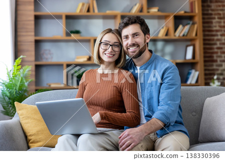 Smiling couple sitting on a sofa in their living room, looking at the viewer while engaging with a laptop, representing connection and modern lifestyle Smiling couple sitting on a sofa in their living room, looking at the viewer while engaging with a laptop, representing connection and modern lifestyle 138330396