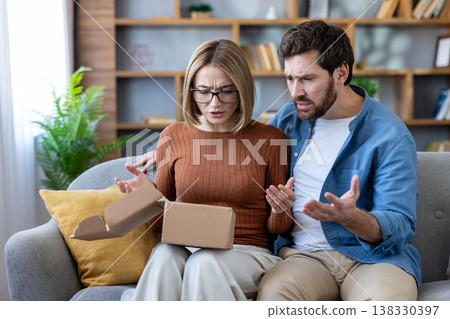 Couple sitting on a couch at home, looking frustrated and expressing disappointment after opening a damaged brown carton box from an online purchase or delivery Couple sitting on a couch at home, looking frustrated and expressing disappointment after opening a damaged brown carton box from an online purchase or delivery 138330397