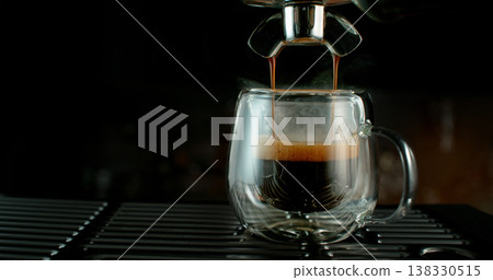 Fresh espresso pouring from coffee machine into glass cup with rich crema and rising steam. Warm cinematic scene capturing preparation of black coffee in cozy atmosphere. 138330515