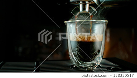 Fresh espresso pouring from coffee machine into glass cup with rich crema and rising steam. Warm cinematic scene capturing preparation of black coffee in cozy atmosphere. 138330517