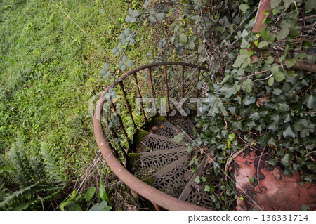 A rusty spiral metal staircase surrounded by dense greenery and overgrown plants in Busaco Park, Portugal. Themes explore decay, abandonment, and the return of nature to man-made structures. A rusty spiral metal staircase surrounded by dense greenery and overgrown plants in Busaco Park, Portugal. Themes explore decay, abandonment, and the return of nature to man-made structures. 138331714