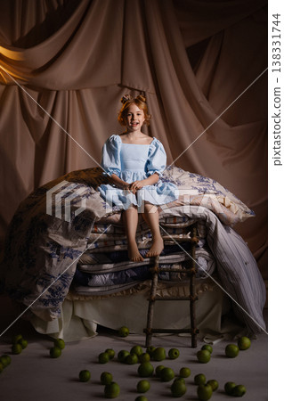 Little girl in blue dress sitting on stacked mattresses with apples, princess concept, studio drapery Little girl in blue dress sitting on stacked mattresses with apples, princess concept, studio drapery 138331744