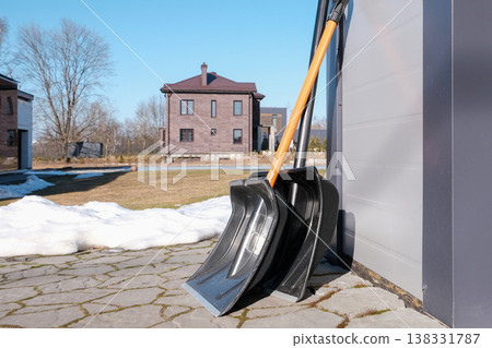 Two snow shovels leaning against a garage with melting snow, ready for winter cleanup. Two snow shovels leaning against a garage with melting snow, ready for winter cleanup. 138331787