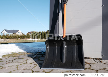 Two snow shovels leaning against a garage with melting snow, ready for winter cleanup. Two snow shovels leaning against a garage with melting snow, ready for winter cleanup. 138331788