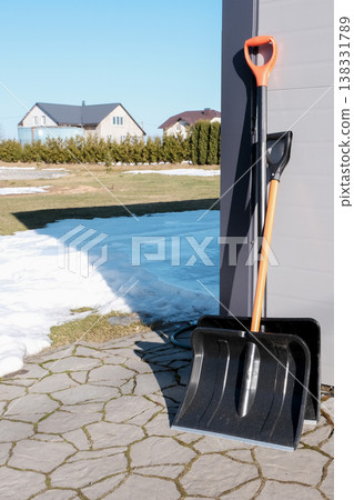 Two snow shovels leaning against a garage with melting snow, ready for winter cleanup. Two snow shovels leaning against a garage with melting snow, ready for winter cleanup. 138331789