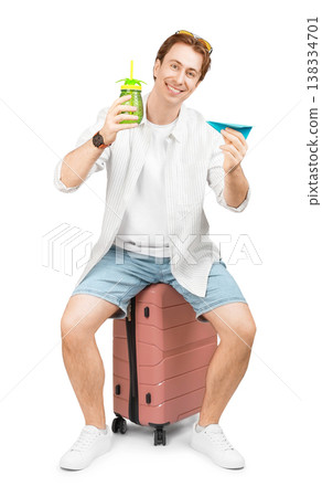 Happy young man ready for summer vacation sitting on a pink suitcase, holding a tropical drink and a blue paper airplane, isolated on a white background. Happy young man ready for summer vacation sitting on a pink suitcase, holding a tropical drink and a blue paper airplane, isolated on a white background. 138334701