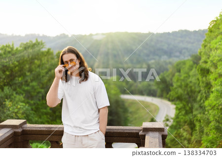 Smiling long-haired man in a white tshirt adjusts sunglasses on a balcony. Scenic background of a winding road, lush green forest, and bright sun rays. Smiling long-haired man in a white tshirt adjusts sunglasses on a balcony. Scenic background of a winding road, lush green forest, and bright sun rays. 138334703