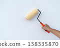 A person's hand holding a paint roller against a clean white textured wall. Home renovation and interior improvement concept with a yellow roller and orange handle on a bright background. 138335870