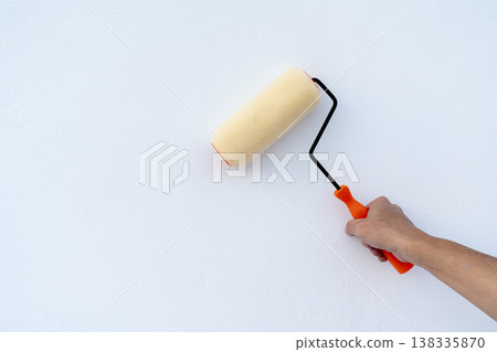 A person's hand holding a paint roller against a clean white textured wall. Home renovation and interior improvement concept with a yellow roller and orange handle on a bright background. 138335870
