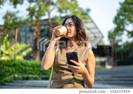 Pretty asian woman holding phone drinking coffee while standing or walking down stairs in park plaza Pretty asian woman holding phone drinking coffee while standing or walking down stairs in park plaza 138337434