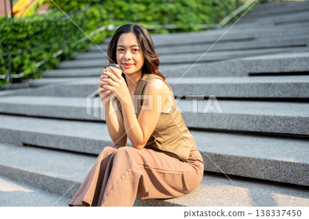 Pretty asian woman holding coffee cup with both hands while sitting on concrete stair under sunlight Pretty asian woman holding coffee cup with both hands while sitting on concrete stair under sunlight 138337450