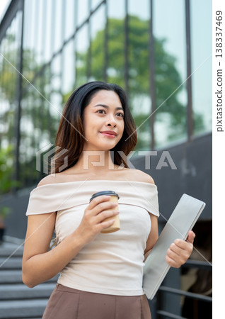 Pretty asian woman holding a laptop and coffee while standing or walking down stairs by the building Pretty asian woman holding a laptop and coffee while standing or walking down stairs by the building 138337469