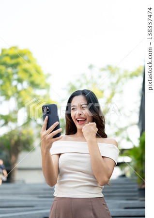Excited asian woman looking at phone making a fist while walking down concrete stairs in park plaza. Excited asian woman looking at phone making a fist while walking down concrete stairs in park plaza. 138337474
