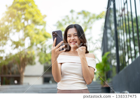 Happy asian woman looking at phone making a fist while walking down concrete stairs by the building. Happy asian woman looking at phone making a fist while walking down concrete stairs by the building. 138337475