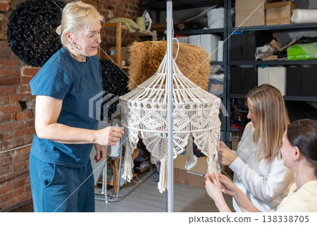 Guiding instructor demonstrating weaving technique while participants assemble decorative chandelier frames with cords in studio. Guiding instructor demonstrating weaving technique while participants assemble decorative chandelier frames with cords in studio. 138338705