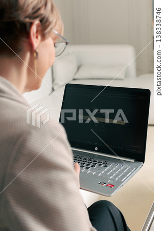 Working focused. Mature businesswoman sits at table uses laptop types on keyboard attentive expression modern interior soft natural light 138338746