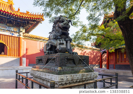 Bronze guardian lion sculpture on pedestal near traditional ornate temple building, red walls golden roofs tree shade in Lama Temple Beijing on sunny day, Beijing, China 138338851
