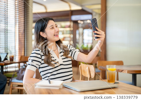 Pretty asian woman holding phone talking on video call aside laptop and notebook at table in cafe. Pretty asian woman holding phone talking on video call aside laptop and notebook at table in cafe. 138339779