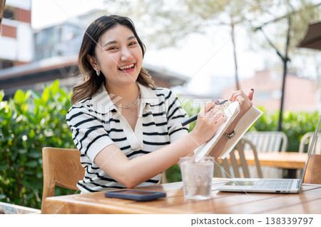 Pretty asian woman holding pen and notebook over laptop while sitting at wooden table in garden cafe Pretty asian woman holding pen and notebook over laptop while sitting at wooden table in garden cafe 138339797