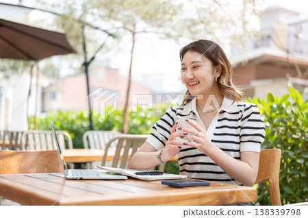 Pretty asian woman talking to video call on laptop aside book while sitting at table in garden cafe. Pretty asian woman talking to video call on laptop aside book while sitting at table in garden cafe. 138339798