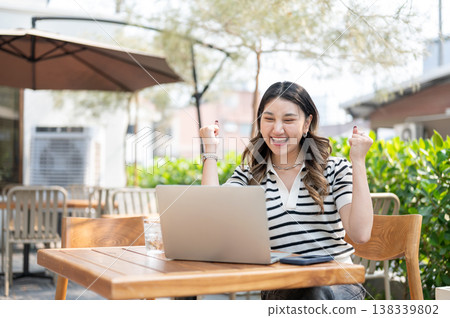 Excited asian woman looking at laptop and making a fist while sitting at wooden table in garden cafe Excited asian woman looking at laptop and making a fist while sitting at wooden table in garden cafe 138339802