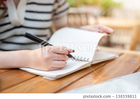 Close up of a woman's hands holding pen and opening a notebook while sitting at wooden table in cafe Close up of a woman's hands holding pen and opening a notebook while sitting at wooden table in cafe 138339811