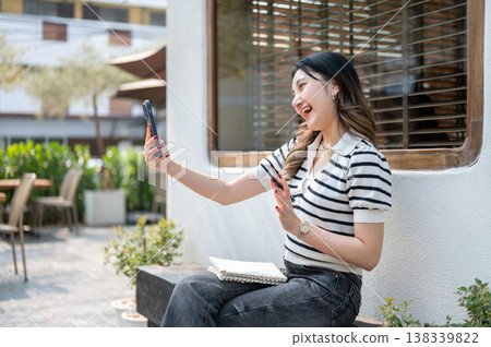 Pretty asian woman holding phone talking on video call while sitting on wooden bench in garden cafe. Pretty asian woman holding phone talking on video call while sitting on wooden bench in garden cafe. 138339822