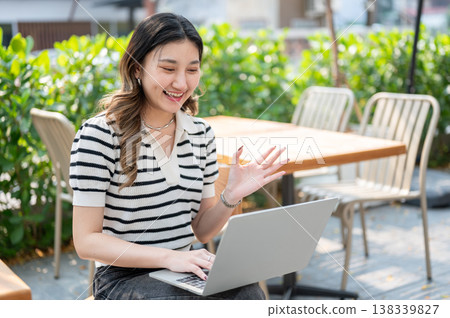 Pretty asian woman talking on video call through laptop while sitting on wooden chair in garden cafe Pretty asian woman talking on video call through laptop while sitting on wooden chair in garden cafe 138339827