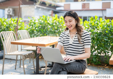 Pretty asian woman looking at laptop holding screen lid while sitting on wooden chair in garden cafe Pretty asian woman looking at laptop holding screen lid while sitting on wooden chair in garden cafe 138339828