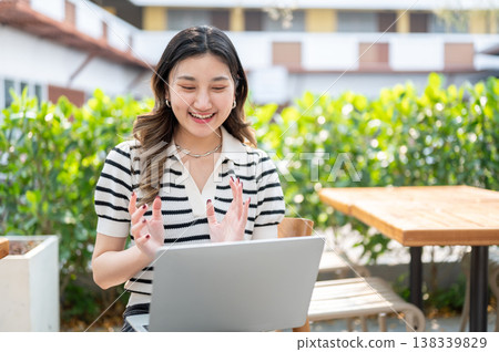 Pretty asian woman talking on video call with a laptop while sitting on wooden chair in garden cafe. Pretty asian woman talking on video call with a laptop while sitting on wooden chair in garden cafe. 138339829
