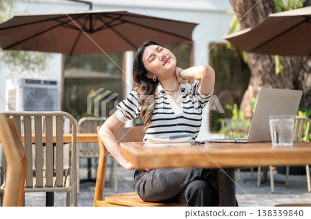 Pretty asian woman holding massaging neck in front of laptop sitting at wooden table in garden cafe. 138339840