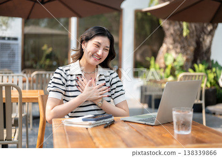 Proud asian woman putting hands over chest looking at laptop sitting at wooden table in garden cafe. Proud asian woman putting hands over chest looking at laptop sitting at wooden table in garden cafe. 138339866